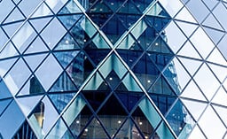  A close-up of a modern skyscraper's glass facade, featuring a repeating geometric pattern of blue-tinted triangular panels and dark metal framing. 