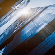 Low-angle shot looking up at modern glass skyscrapers with bright sunlight bursting between the buildings.