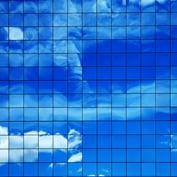 A grid of square, blue glass windows on a modern building facade reflecting a bright blue sky with white clouds.
