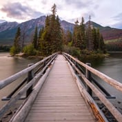 A balanced, symmetrical view of a wooden bridge leads across a calm lake to a small, pine-covered island, with large mountains rising in the background.