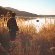 A person stands in a field of tall grass looking out over a calm lake at sunrise.