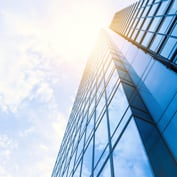 A modern glass office building towers against a partly cloudy sky, with sunlight gleaming off its blue windows.