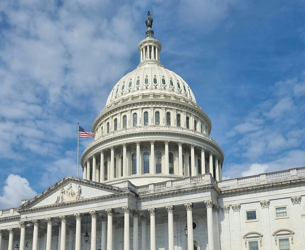 A close up image of the US Capitol building against a blue sky with clouds. An American flag flies nearby.