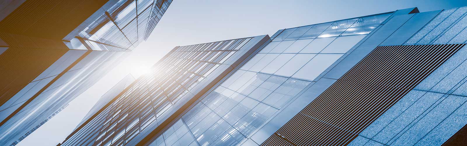 Low-angle shot looking up at modern glass skyscrapers with bright sunlight bursting between the buildings.