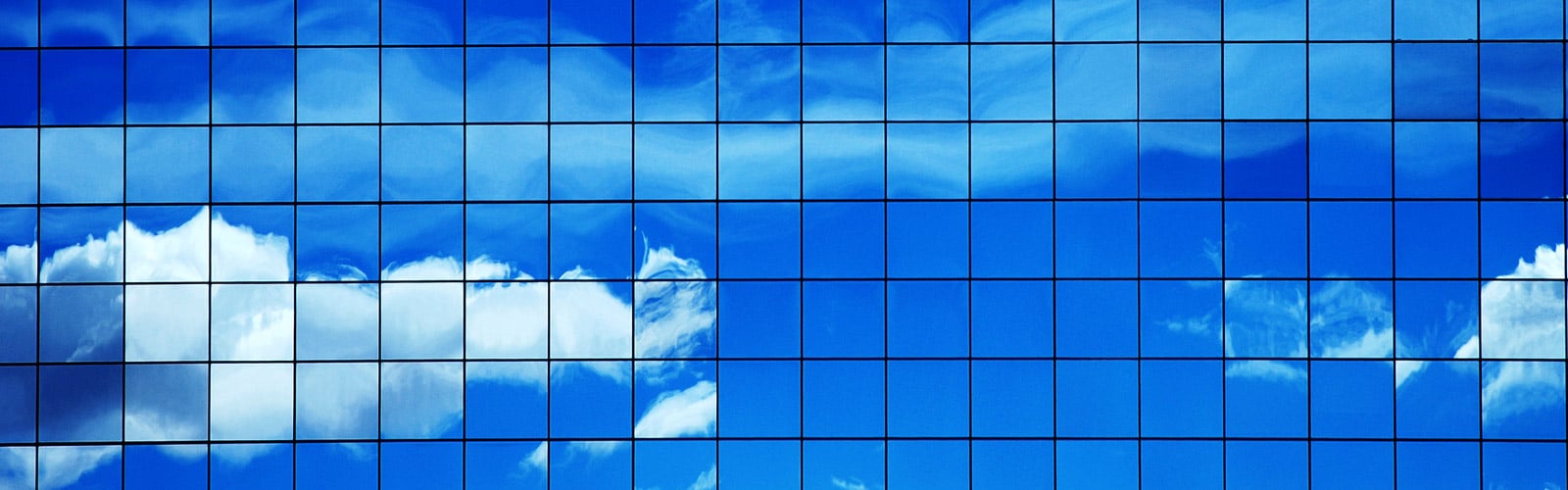 A grid of square, blue glass windows on a modern building facade reflecting a bright blue sky with white clouds.